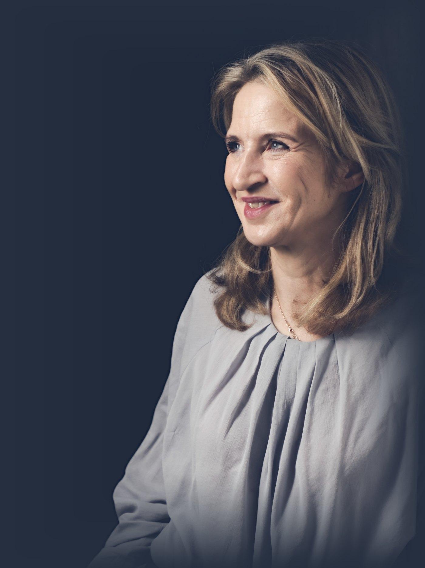 Porträt von Katrin Funke, an eine Wand gelehnt, den Blick zur Seite gerichtet; warme gedämpfte Farben, der dunkle Hintergrund läuft in die Seite aus.