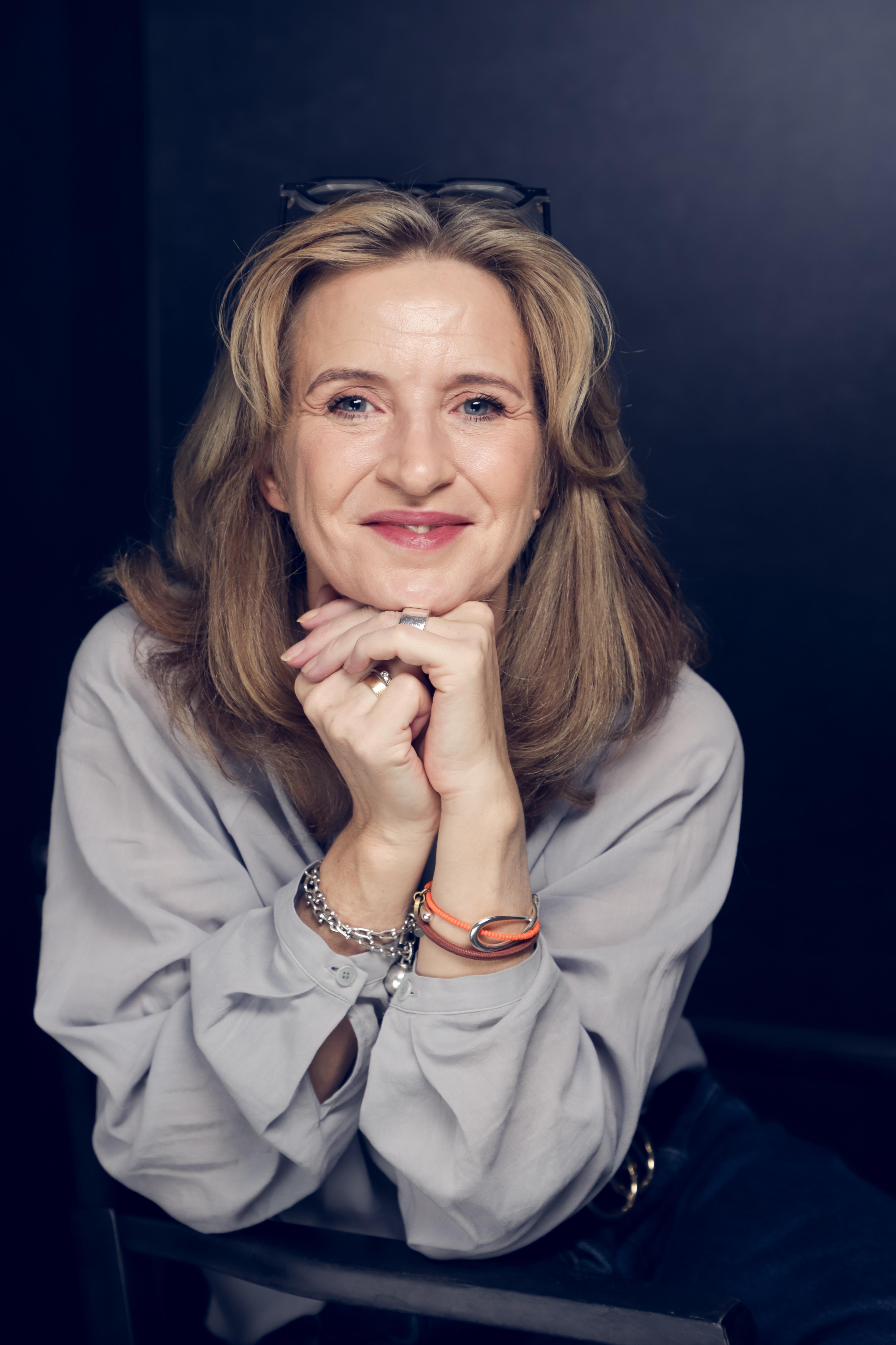Katrin Funke, sitzend, das Kinn auf die Hände gestützt, mit warmem Lächeln direkt in die Kamera.