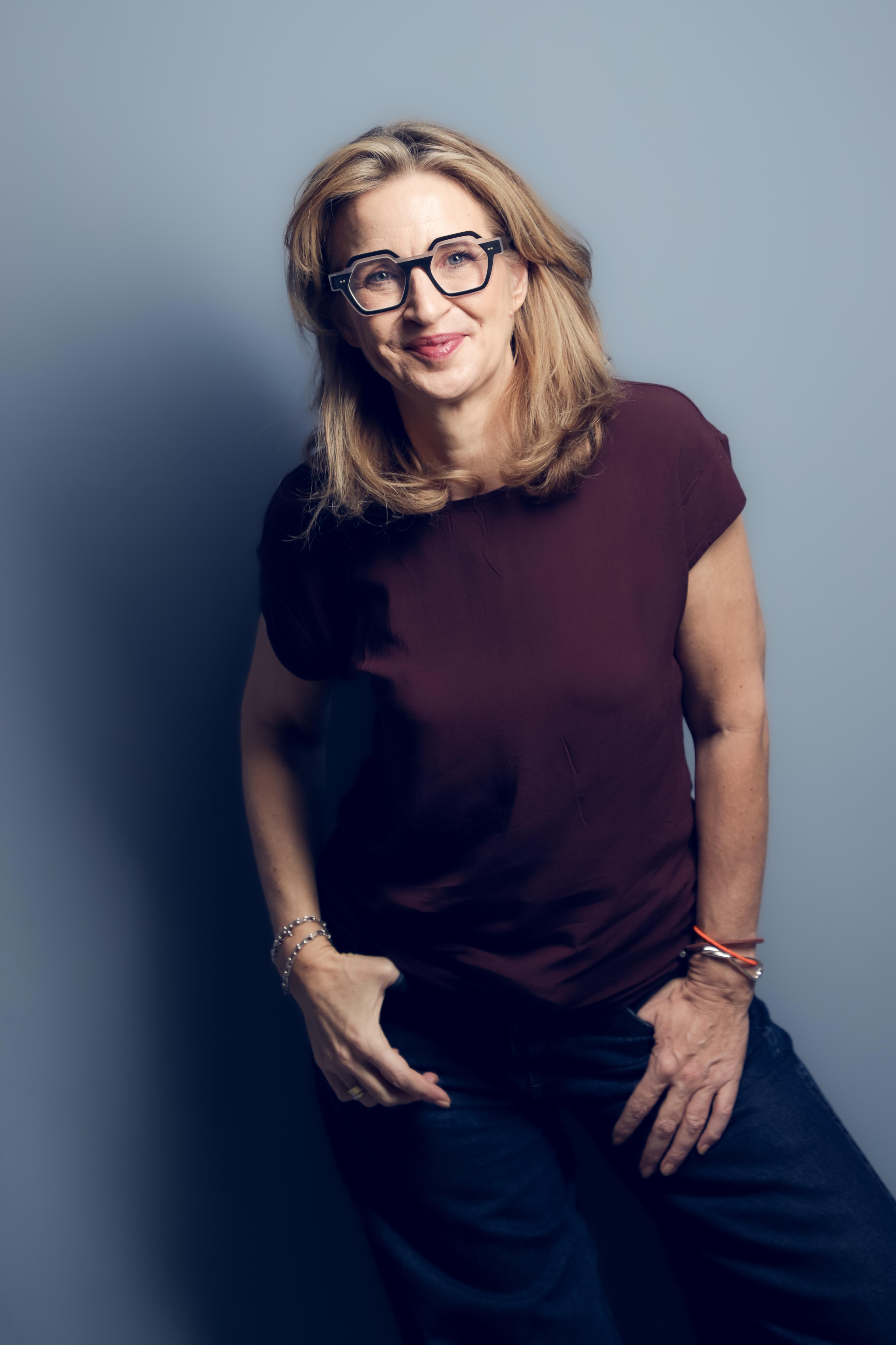 Katrin Funke im Stehen, mit markanter Brille und kastanienrotem Oberteil, selbstbewusst lächelnd.