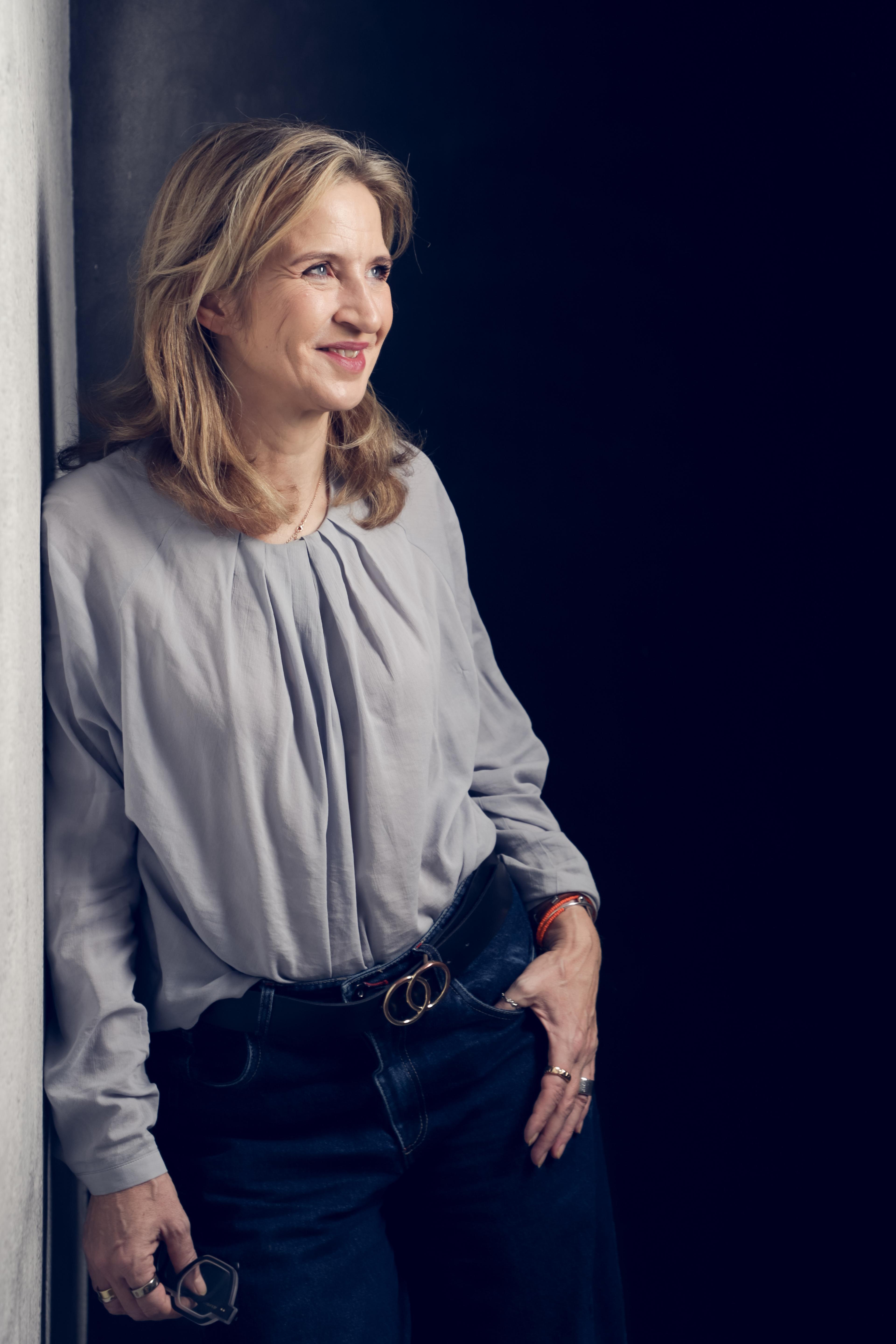 Katrin Funke an eine Wand gelehnt, den Blick zur Seite gerichtet, in grauer Bluse vor dunklem Hintergrund.